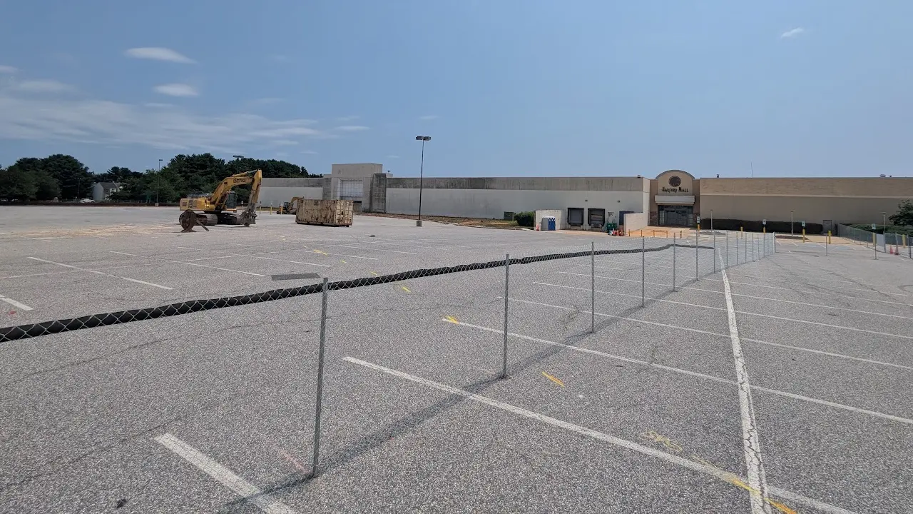 Fenced former Macy's work zone at Harford Mall with an excavator and the exterior building still standing