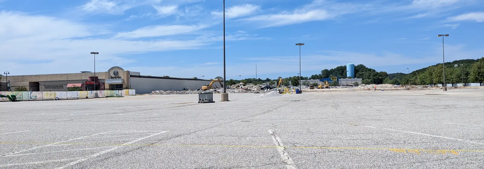 Wide east-side view across the former Macy's footprint at Harford Mall, now mostly open ground and rubble, with the retained mall at left and demolition equipment at center
